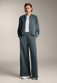 Giacca leggera zip-up color turchese con cappuccio, abbinata a pantaloni larghi a coste color turchese. Sotto indossa una maglietta bianca aderente. Presenta tasche.