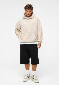 Beige oversized hoodie met een voorzak en zwart logo, gecombineerd met zwarte shorts en witte sokken, afgerond met beige en zwarte sneakers.