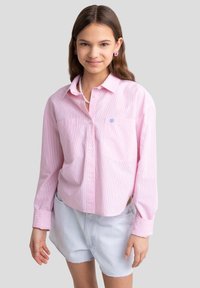 Camicia button-up a righe rosa chiaro con maniche lunghe, due tasche sul petto e design ricamato blu. Abbinata a pantaloni corti di jeans azzurri.