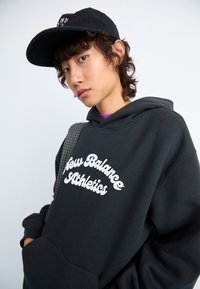 Jeune personne portant un hoodie noir New Balance Athletics et une casquette noire, regardant l'appareil photo avec une main dans la poche du hoodie, sur fond uni.
