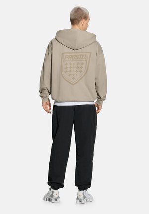 Mężczyzna z krótkimi włosami, zwrócony tyłem, ubrany w beżową bluzę z kapturem z logo "PROSTO", czarne spodnie i białe trampki na białym tle.