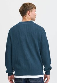 Teksturl blå genser med ribbestrikket mønster, rund hals og droppe skuldre. Båret over en hvit skjorte og paret med mørke jeans.