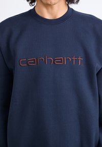 Navy blue sweatshirt made of soft fabric, featuring raised brown "carhartt" text logo across the chest, with a crew neck design.