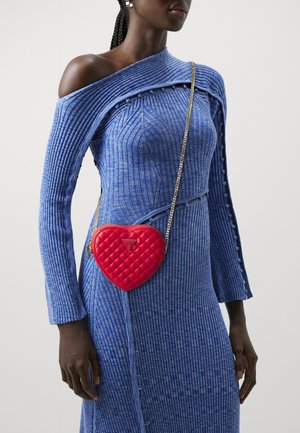 Bolso bandolera acolchado en forma de corazón rojo con cadena, llevado sobre un vestido azul de canalé con botones decorativos en las mangas.