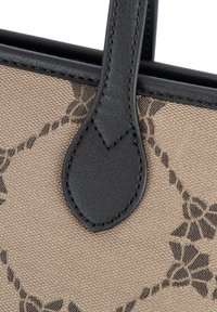 Czarny skórzany uchwyt w kształcie pętli przymocowany do beżowej tkaniny plecionej z czarnym wzorem kwiatowym w łańcuch.