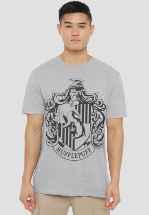 Junger Mann trägt ein hellgraues T-Shirt mit großem schwarzem Hufflepuff-Wappen vorne, steht vor einfarbigem Hintergrund.