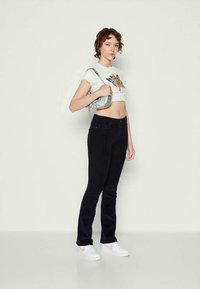 Ung kvinne med kort krøllete hår som har på seg en hvit sommerfugl crop top, svarte bukser, hvite joggesko og holder en liten skulderveske.