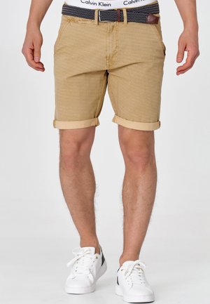 Shorts beiges à motifs avec des ourlets retroussés, une ceinture à motifs bleu marine et blanc sur la taille, associés à des baskets blanches.