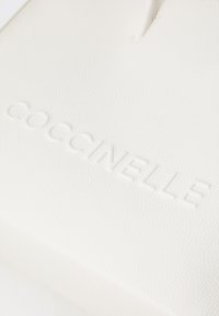 Sac en cuir blanc avec logo "Coccinelle" en relief, texture lisse, forme rectangulaire et design minimaliste.