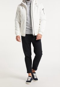 Witte gewatteerde jas met rits en trekkoordkap, grijze trui eronder, zwarte broek en marineblauwe sneakers met witte veter.