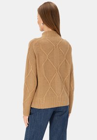 Beżowy sweter z dzianiny z wysokim kołnierzem i teksturą w diamentowy wzór. Posiada prążkowany dół i rękawy, stylizowany z niebieskimi dżinsami.