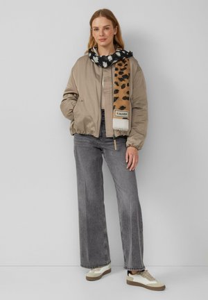 Beige bomberjack, grijze wijde jeans en een zwarte sjaal met witte patronen en accenten van luipaardprint. Sneakers met een beige en witte kleurencombinatie.