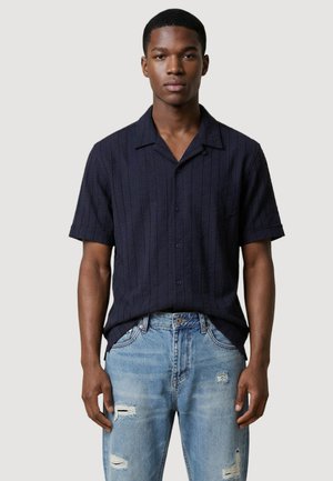 Giovane uomo con camicia scura a maniche corte e jeans chiari strappati, in piedi davanti a uno sfondo chiaro e semplice.