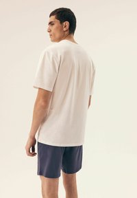 Biała bawełniana koszulka z krótkim rękawem, widok z tyłu, ukazująca luźny krój; zestawiona z granatowymi szortami, o prostym wzorze i gładkiej teksturze.