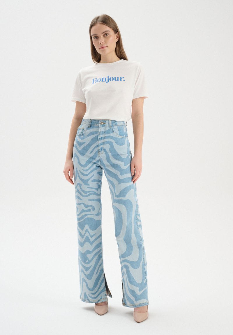 Camiseta blanca con "Bonjour." impresa en azul. Pantalones vaqueros de un azul claro con un patrón ondulado y aberturas laterales; ambos artículos tienen una textura suave.