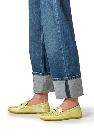 Scarpe gialle a fantasia senza lacci con una texture liscia e accessori minimi, abbinate a pantaloni di jeans blu risvoltati.