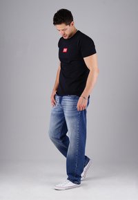 Mann in einem schwarzen T-Shirt mit rotem Logo, blauen Jeans und weißen Sneakers, steht da und schaut auf einen grauen Hintergrund.