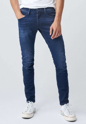Man draagt donkerblauwe slim-fit jeans met subtiel vervagen en witte sneakers, staande met één hand in de zak.