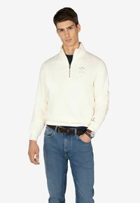 Pullover zip-up di colore crema chiaro realizzato in tessuto morbido, con colletto alto e dettagli ricamati minimi. Abbinato a jeans blu e una cintura intrecciata.