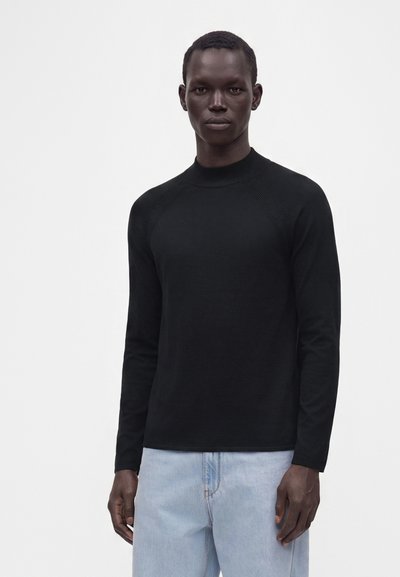 Personne grande à la peau foncée portant un haut noir ajusté à manches longues et un jean bleu clair, debout devant un fond clair uni.
