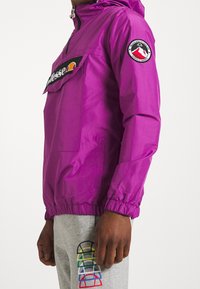 Fioletowa wiatrówka z przednią kieszenią, elastycznymi mankietami i akcentami z logo. Połączona z szarymi dresami z kolorowymi, graficznymi detalami.
