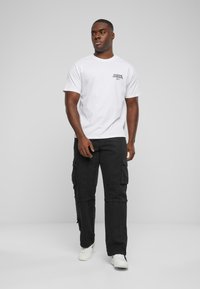 Hvid bomulds t-shirt med et sort grafisk logo på venstre bryst, parret med sorte cargo-bukser med lommer, båret med hvide sneakers.