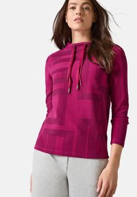 Fuchsia-langärmliger Hoodie mit geometrischem schwarzen Linienmuster, verstellbarem Kordelzug und weichem Stoff. Kombiniert mit hellgrauen Hosen.