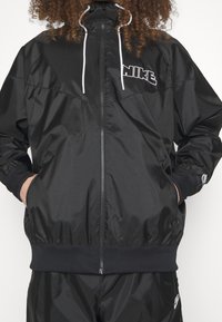 Nike svart vindtät jacka med dragkedja fram, elastiska ärmslut, ståkrage och en vit logotyp. Tillverkad av smidig, lättvävd textil.