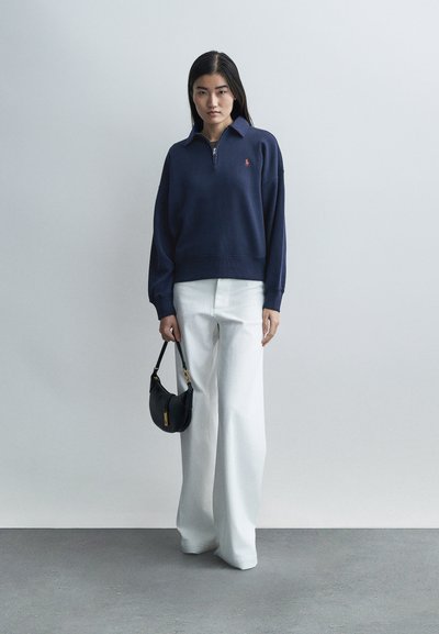 Γυναίκα με μακριά μαύρα μαλλιά που φοράει μπλε πουλόβερ και άσπρο φαρδύ παντελόνι, κρατώντας μια μικρή μαύρη τσάντα, στέκεται μπροστά σε απλό ανοιχτό φόντο.