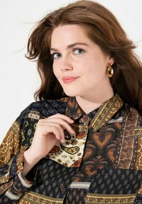 Camisa de botones multicolor con patrones de paisley, texturas variadas y tonos de verde, marrón y crema. Botones marrones acentúan el diseño.