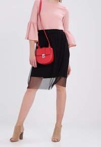 Žena v růžovém topu s širokými rukávy, černé průhledné sukni, béžových otevřených lodičkách a s červenou kabelkou přes rameno.