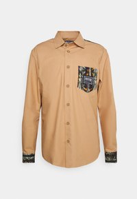 Ljusbrun långärmad skjorta med en mönstrad ficka. Har knappstängning och kontrasterande mönstrade manschetter. Tillverkad av ett mjukt tyg.