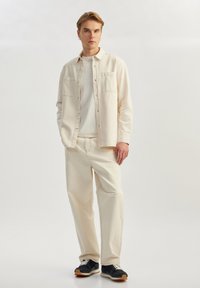 Beige skjorte med knapper og to brystlommer over en hvid t-shirt, parret med lyse beige bukser og sorte sneakers med hvid sål.