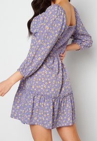 Lila Blumenkleid mit Puffärmeln, gerafftem Oberteil und schwingendem Saum. Es zeigt winzige gelbe Blumen auf einem leichten, strukturierten Stoff.