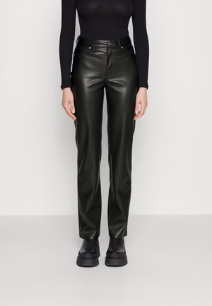 Mujer con top ajustado de manga larga negro, pantalones de cuero sintético negro de talle alto y botas negras gruesas con plataforma, de pie sobre un fondo liso.