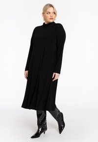 Schwarzes langärmliges Kleid mit hohem Kragen, fließender Silhouette und glatter Textur, kombiniert mit knielangen glänzenden schwarzen Stiefeln.