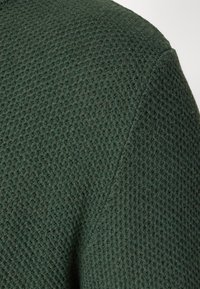 Dark green textured knit fabric with a waffle pattern, showing a close-up of the shoulder seam and overall fabric construction.