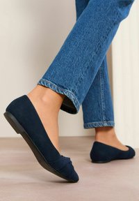 Ballerines en daim bleu marine avec un détail torsadé à l'avant et une semelle en caoutchouc noir basse, associées à un jean court bleu clair.