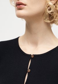 Γυναίκα που φοράει μαύρη ριμπ πλεκτή μπλούζα με καφέ κουμπιά και μεγάλο διαφανές μπεζ κρίκο σκουλαρίκι, εμφανιζόμενη από το λαιμό έως το πηγούνι.