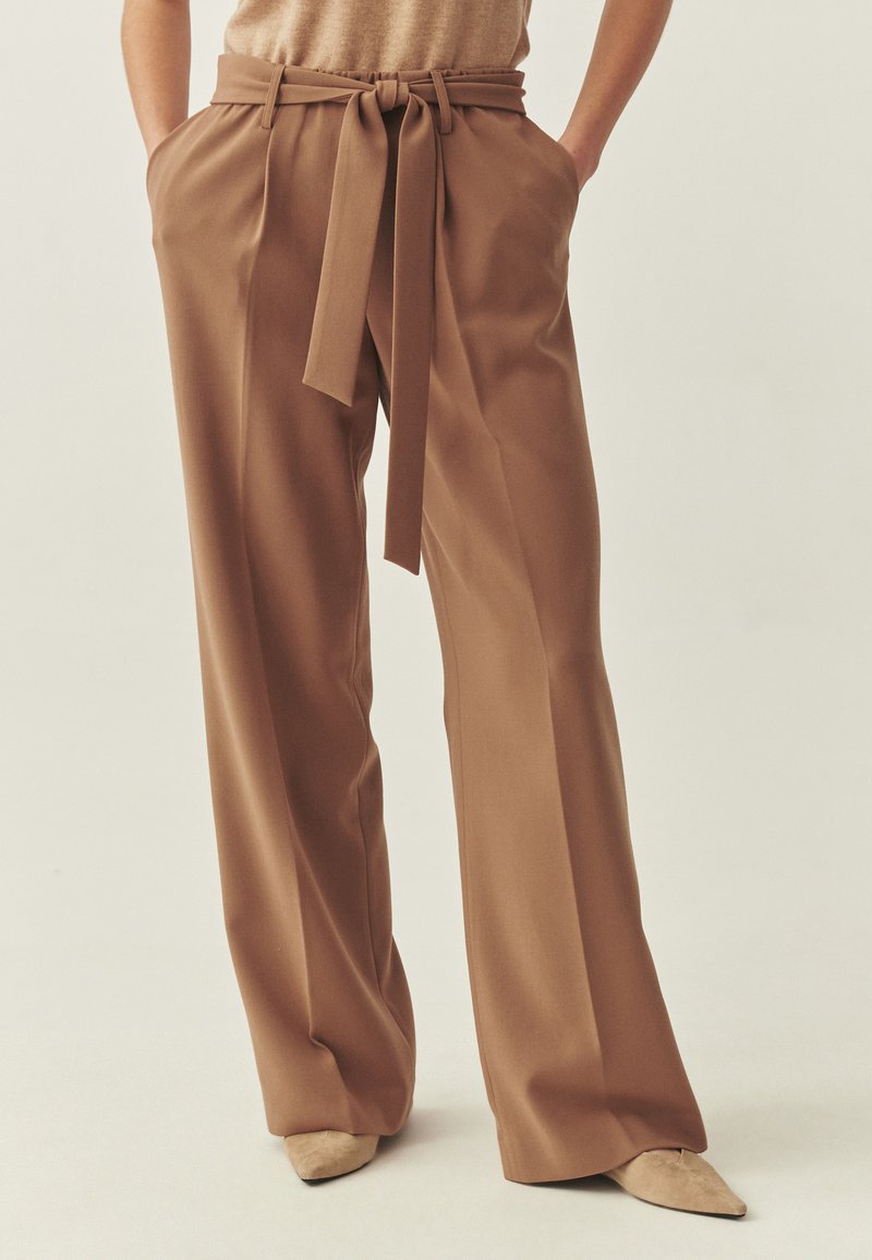 Pantalon marron à jambes larges avec ceinture assortie, présentant une taille haute et deux poches latérales. Le tissu semble lisse avec une coupe élégante.
