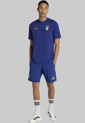 Hombre vistiendo camiseta y pantalones cortos de fútbol de Italia en azul con logos dorados, calcetines blancos con rayas negras y zapatillas deportivas blancas sobre fondo gris.