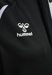 Gros plan sur un tissu noir avec le logo blanc de Hummel et un texte, présentant un dessin stylisé d'une abeille et une partie d'une fermeture éclair.