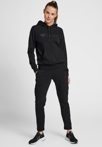 Czarna bluza z kapturem, z kieszenią z przodu, sznurkiem do ściągania i wytłoczonym logo. Zestawiona z dopasowanymi czarnymi dresami, z ściągaczami na rękawach i kieszeniami.