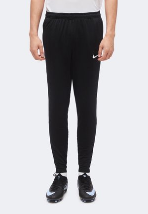 Personne portant un pantalon de sport noir Nike et des crampons de football noirs, debout devant un fond clair uni.