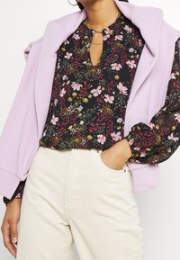 Blouse noire à motifs floraux roses, jaunes et verts ; encolure avec ouverture en forme de goutte. Pull violet clair posé sur les épaules. Pantalon en velours côtelé crème.