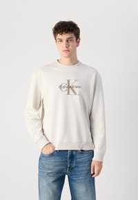 Svijetlosmeđi dukserica s okruglim izrezom i rebrastim manžetama, koja središnjem dijelu ima zlatni logo "calvinKlein". Uparena s plavim trapericama.