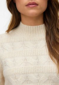 Sweter w kolorze kremowym, wykonany na drutach, z wysokim ściągaczem i wzorem w warkocze, o miękkiej fakturze i otwartym splocie.