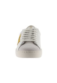 Baskets blanches avec un bout en suède gris, des lacets plats et un logo doré sur le côté. Elles présentent une semelle blanche texturée et un design minimaliste.