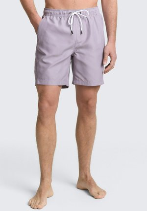 Man draagt lichtpaarse zwemshorts met witte trekkoord, staat blootsvoets tegen een effen achtergrond.