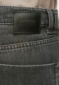 Šedé džínové džíny s černou koženou etiketu s vyraženým nápisem "DRYKORN", viditelným prošíváním na kapsách a hladkou texturou.